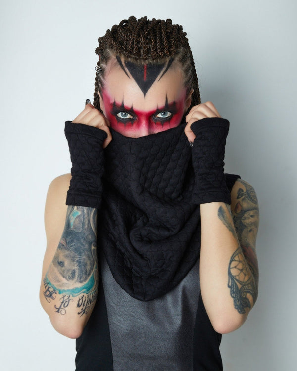 A person covering their face with a black circle scarf, wearing a black tank top, with visible tattoos on the arms.